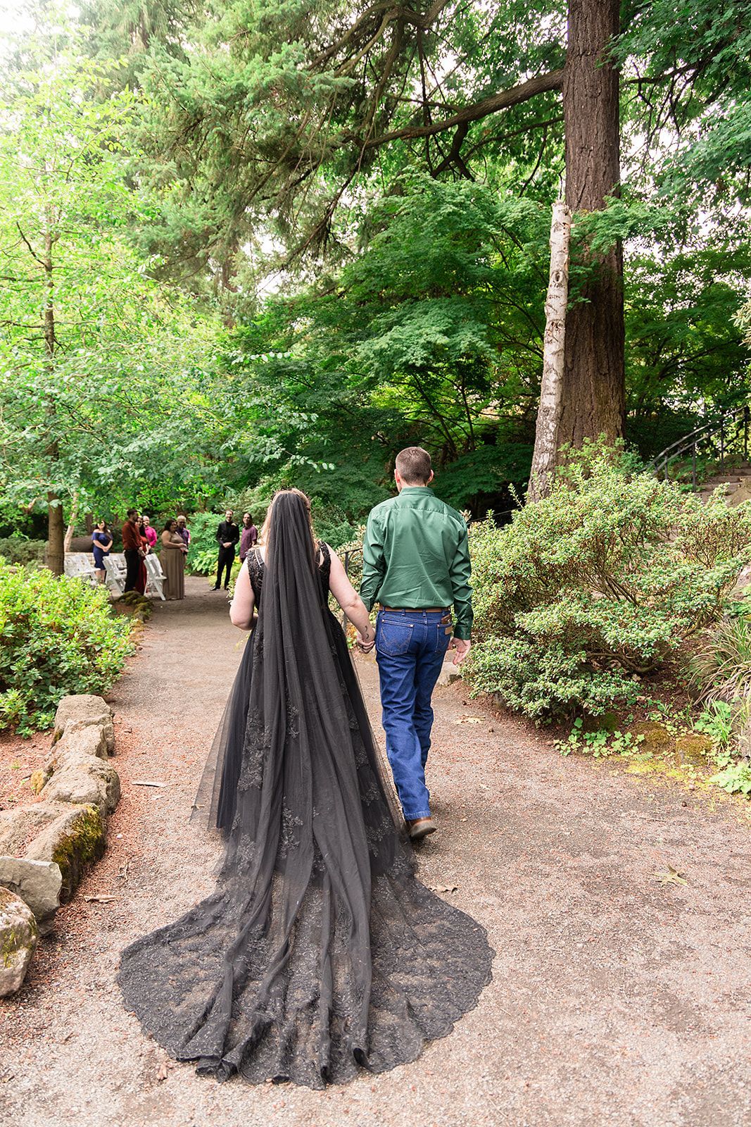 A woman in a black dress and veil is walking down a path with a man