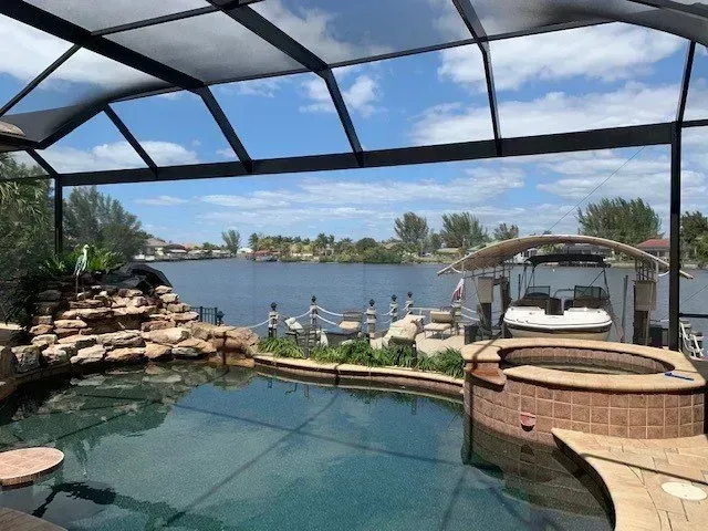 Poolside view of a lake with a covered patio, boat dock, and boats. Blue sky and water, rock waterfall.