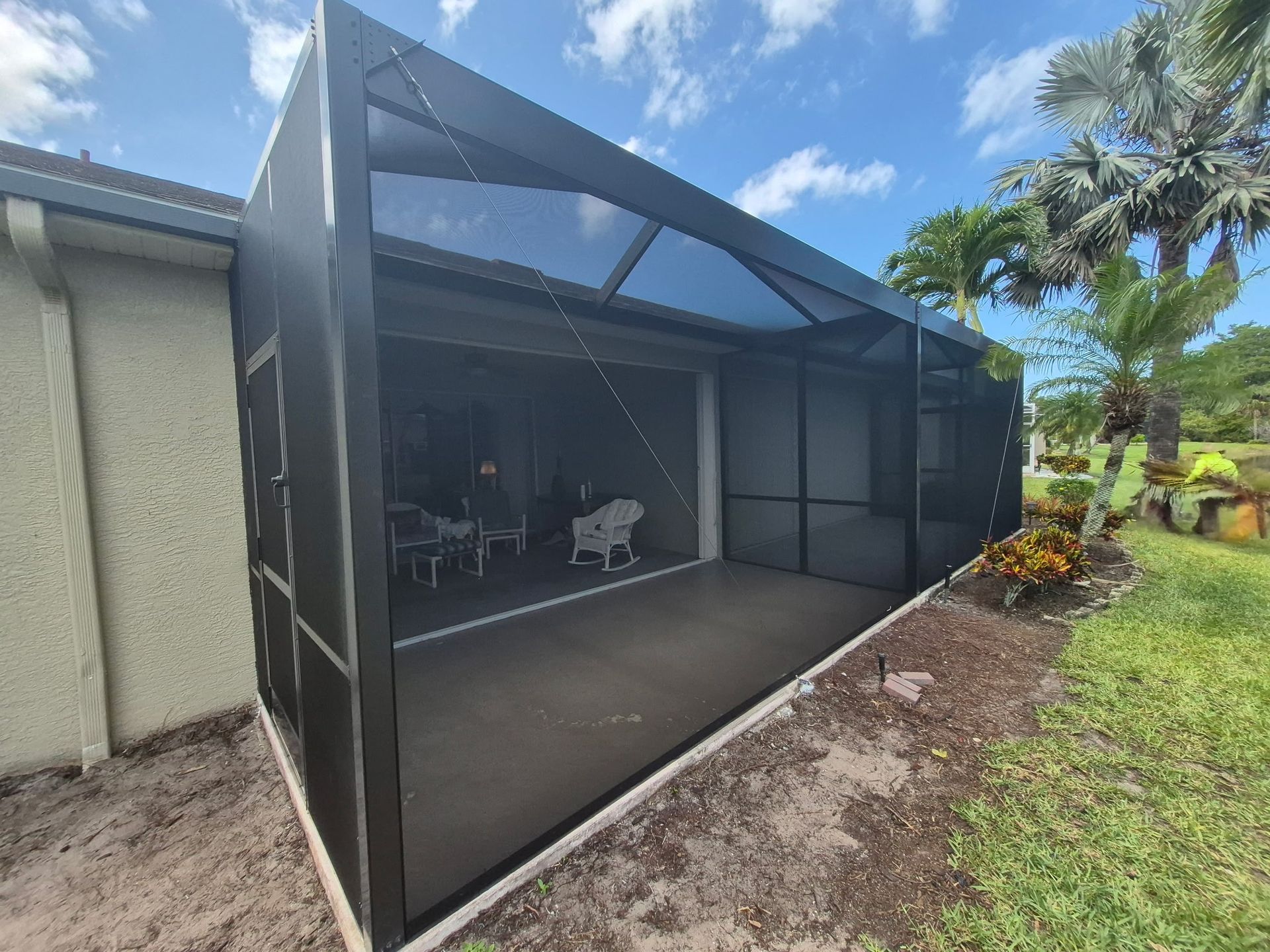 Screened-in patio extension. Black mesh walls and roof, attached to a house. Sunny day with greenery in the background.