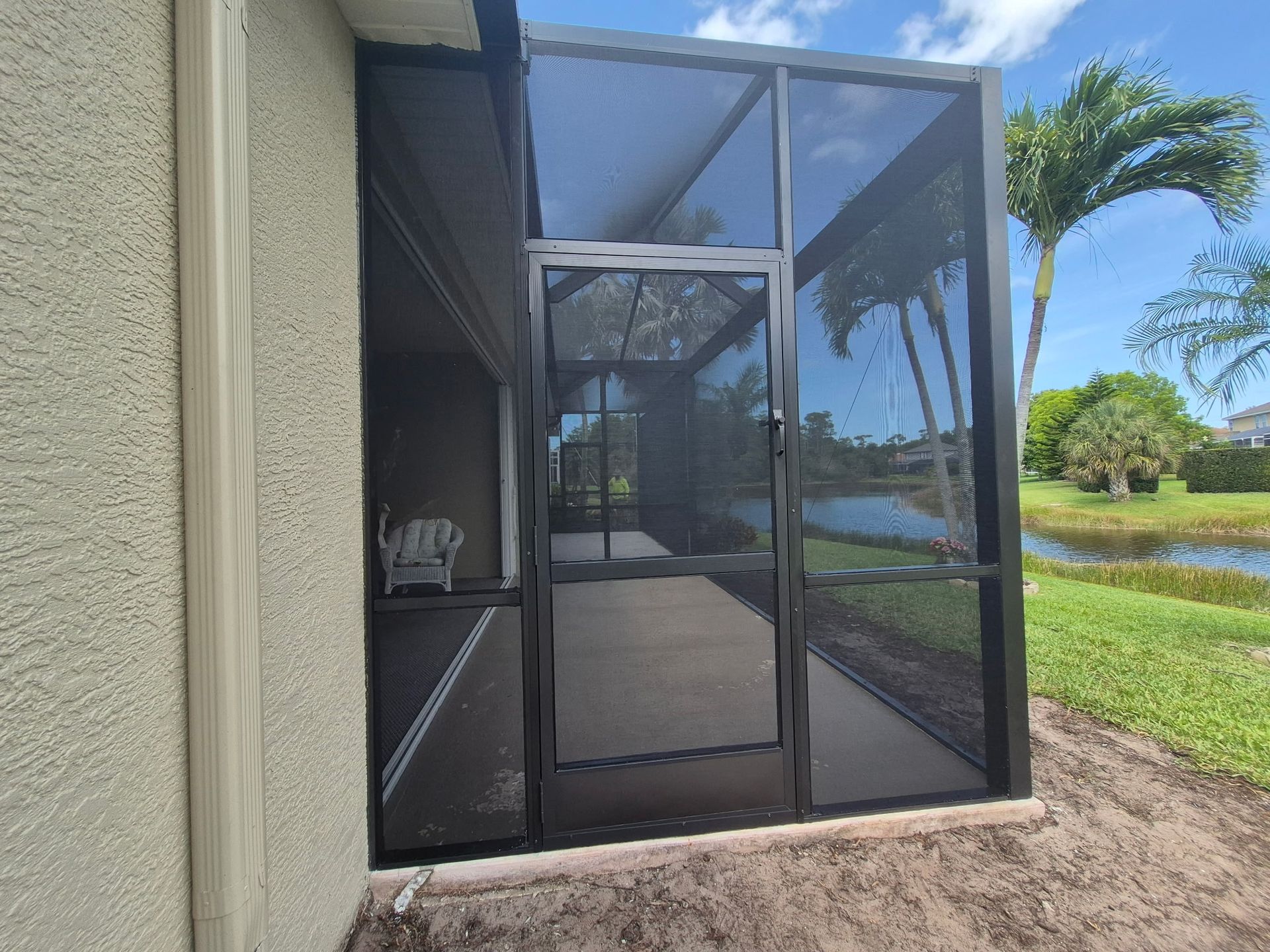 Screened-in porch attached to a stucco building with a door, overlooking a grassy area and water.