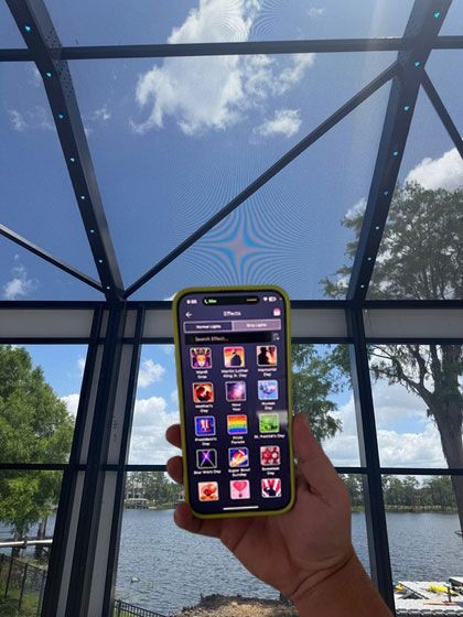 Hand holding a smartphone with app icons, framed against a glass wall and blue sky over water.