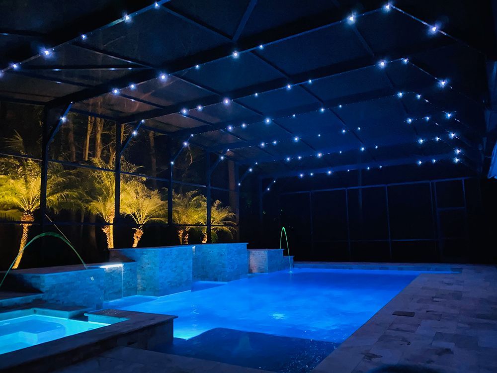 Blue-lit nighttime pool with deck chairs and a covered patio, surrounded by trees and dark fencing.