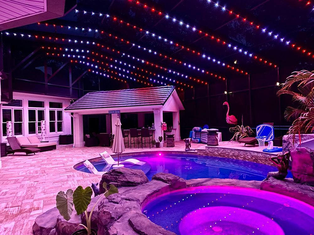 Purple-lit pool patio with string lights, lounge chairs, and a small cabana at night.