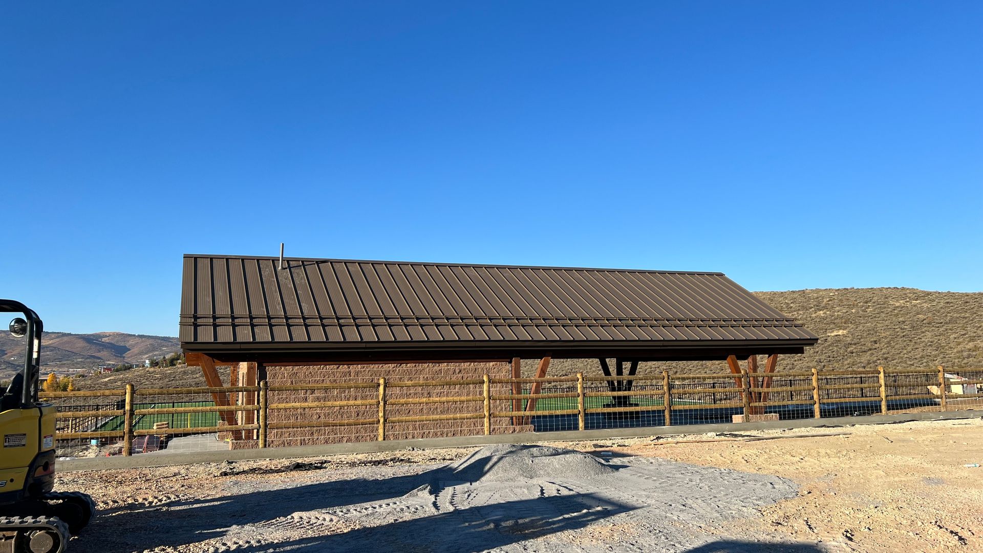 Brown roofed shelter with wooden fence against a hilly backdrop under a clear blue sky.