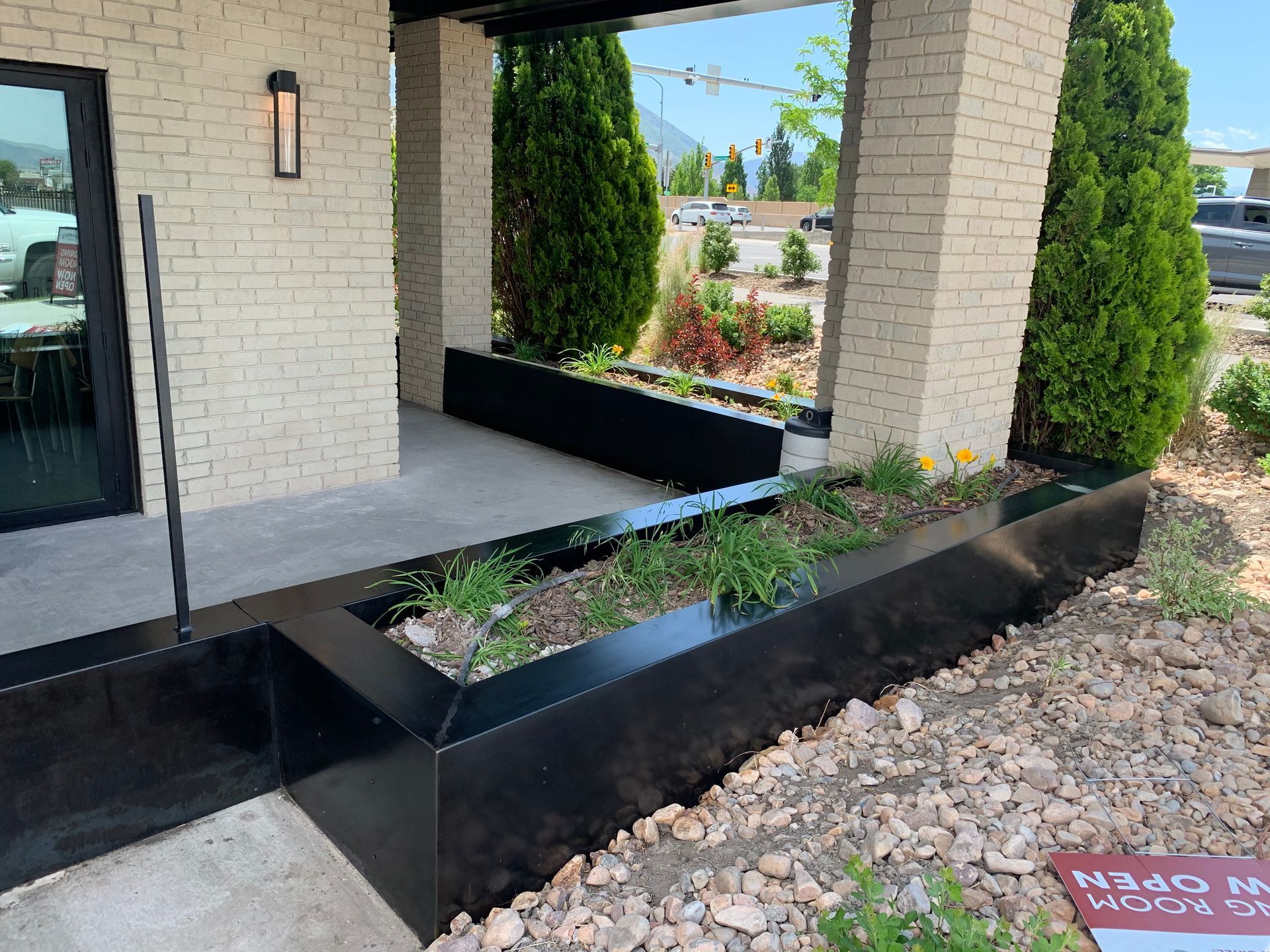Black metal planters with greenery and stone in front of a building with brick pillars.