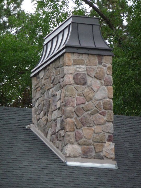 Stone chimney on a dark shingled roof with a black metal chimney cap.