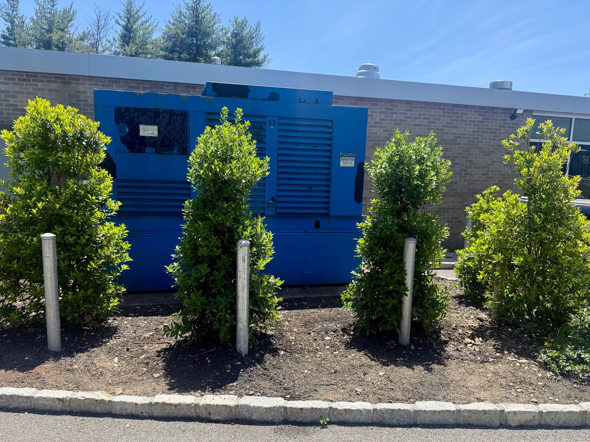 Blue utility box behind neatly planted shrubs and white posts along a curb in bright sunlight