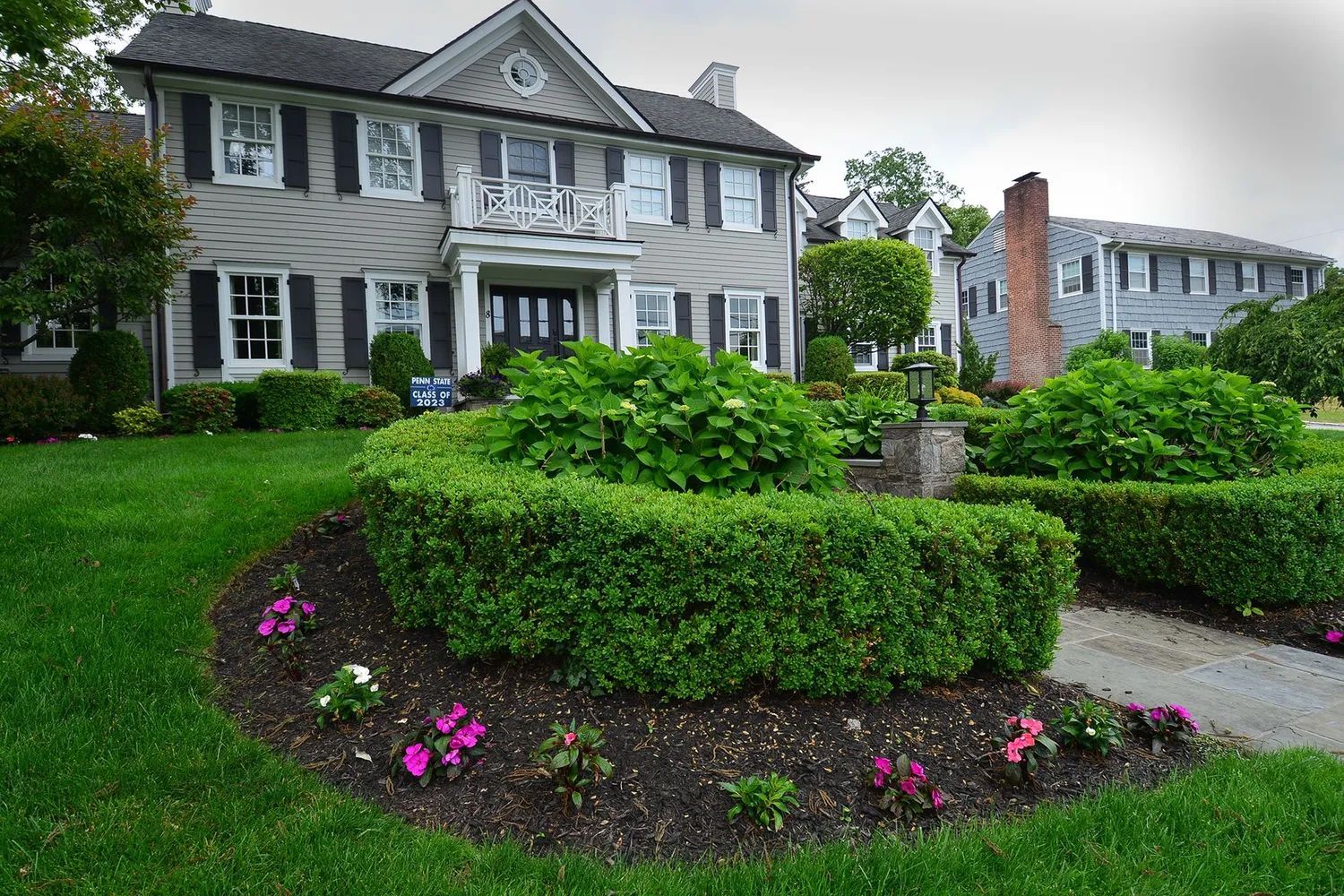 Two-story grey house with green lawn, shrubs, and flower beds.