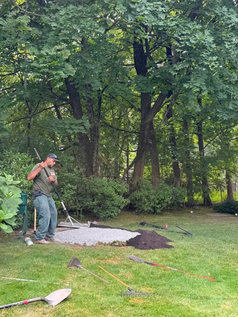 Person shoveling gravel in a grassy backyard beside trees and garden tools