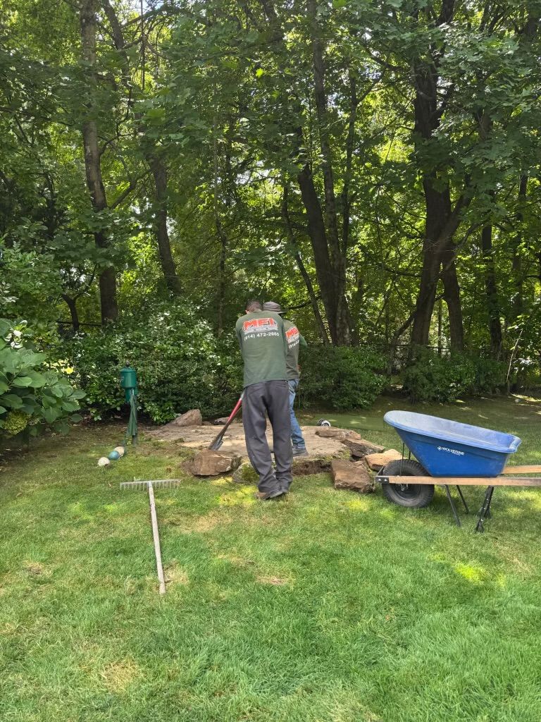 Person spreading soil in a backyard garden beside a blue wheelbarrow and green trees.