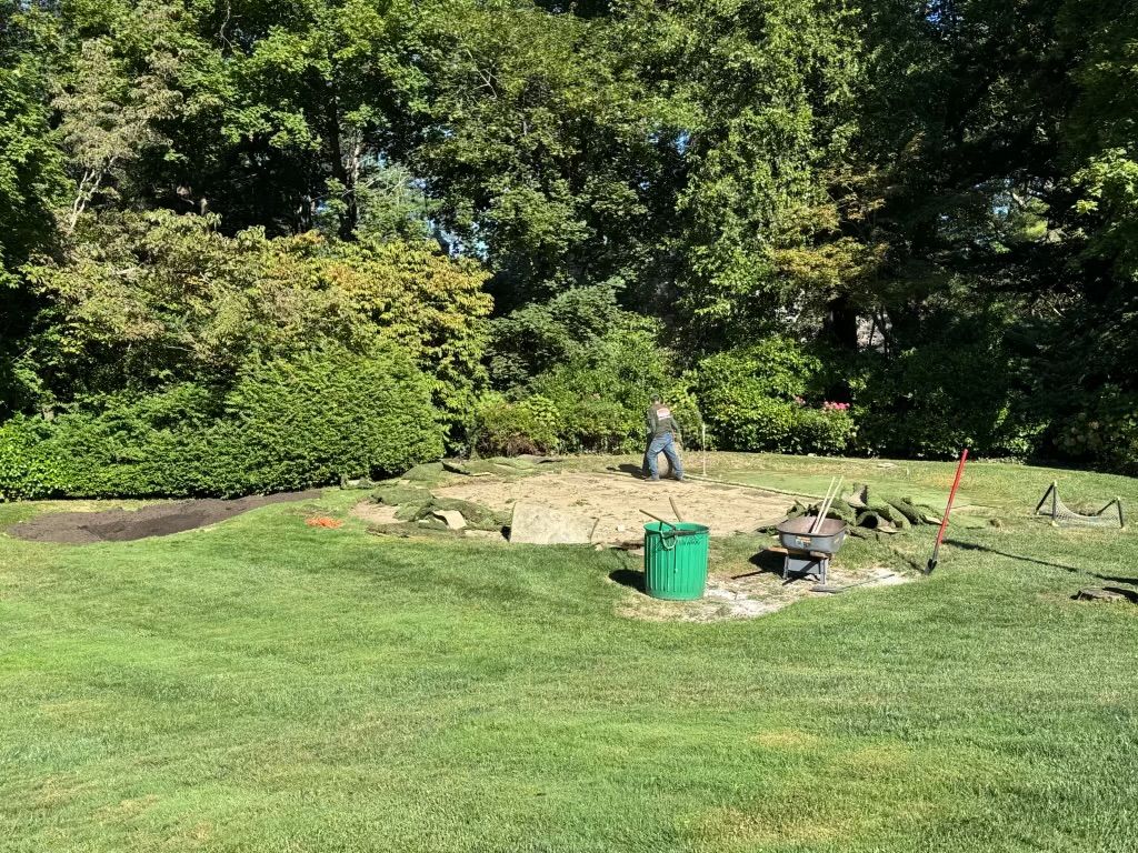 Person standing in a grassy yard near a stone fire pit, with trees and shrubs in the background
