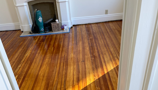 Hardwood floors in sunlight, leading to a fireplace with white trim and items in front.