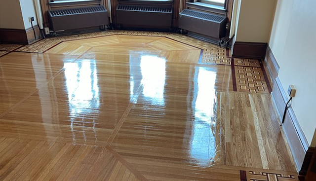 Shiny, newly-refinished wooden floor with intricate geometric patterns. Windows in the background reflect sunlight.