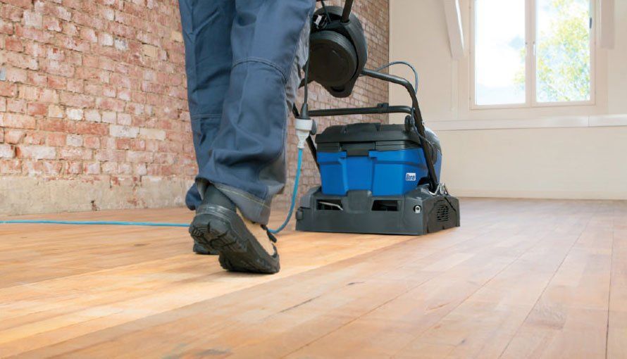 Person sanding a wooden floor with a blue and black machine in a room with a brick wall and a window.