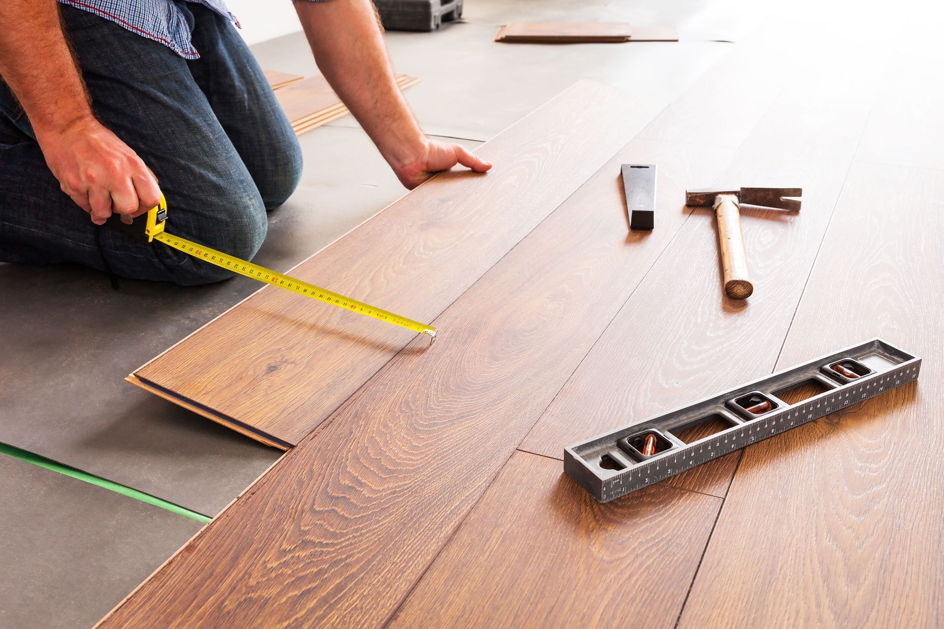 Person kneeling, measuring plank for installing wood flooring. Tools and boards visible.