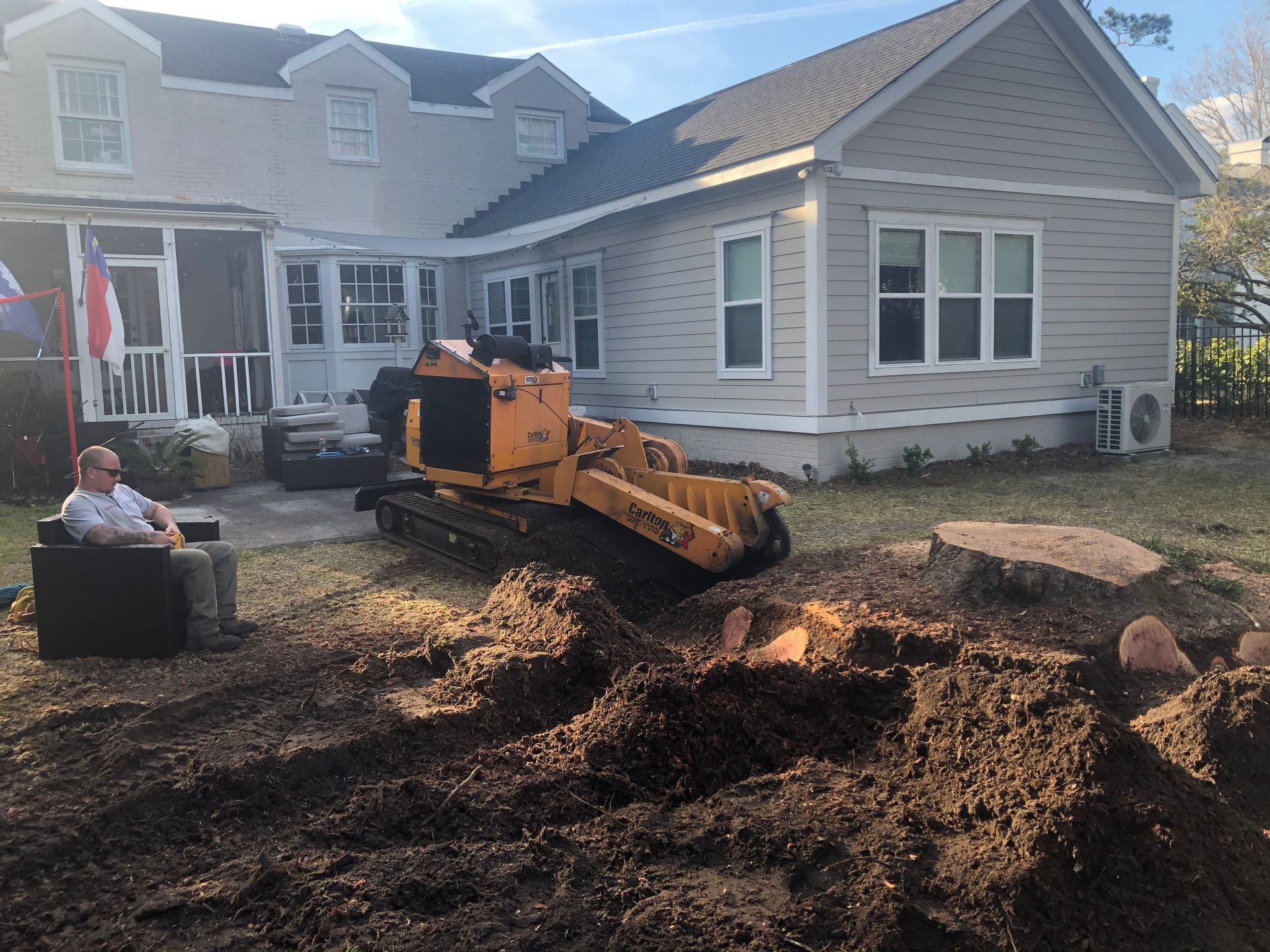 A man is sitting in a chair in front of a house next to a stump grinder.