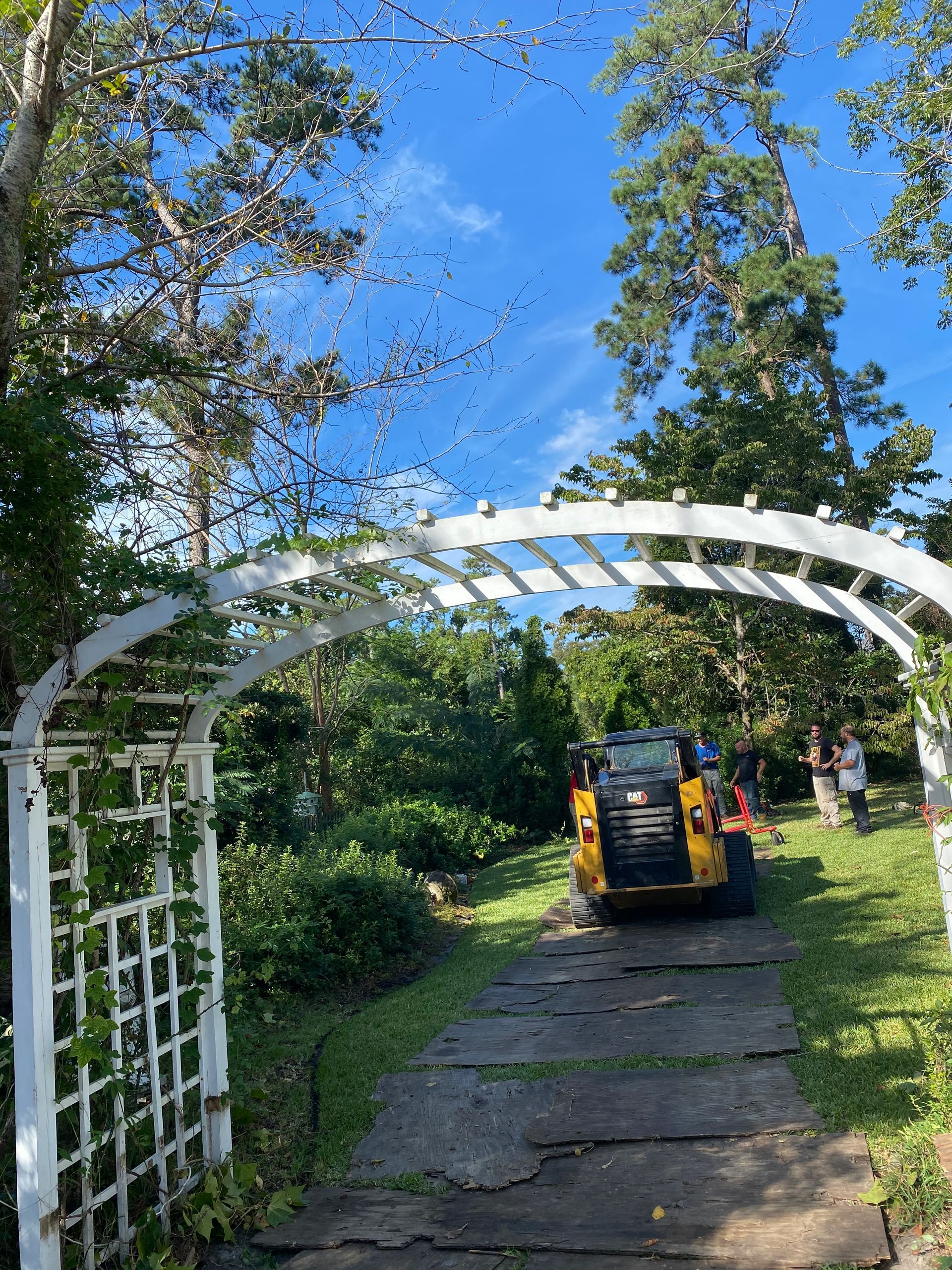 A yellow tractor is driving under a white archway in a garden.