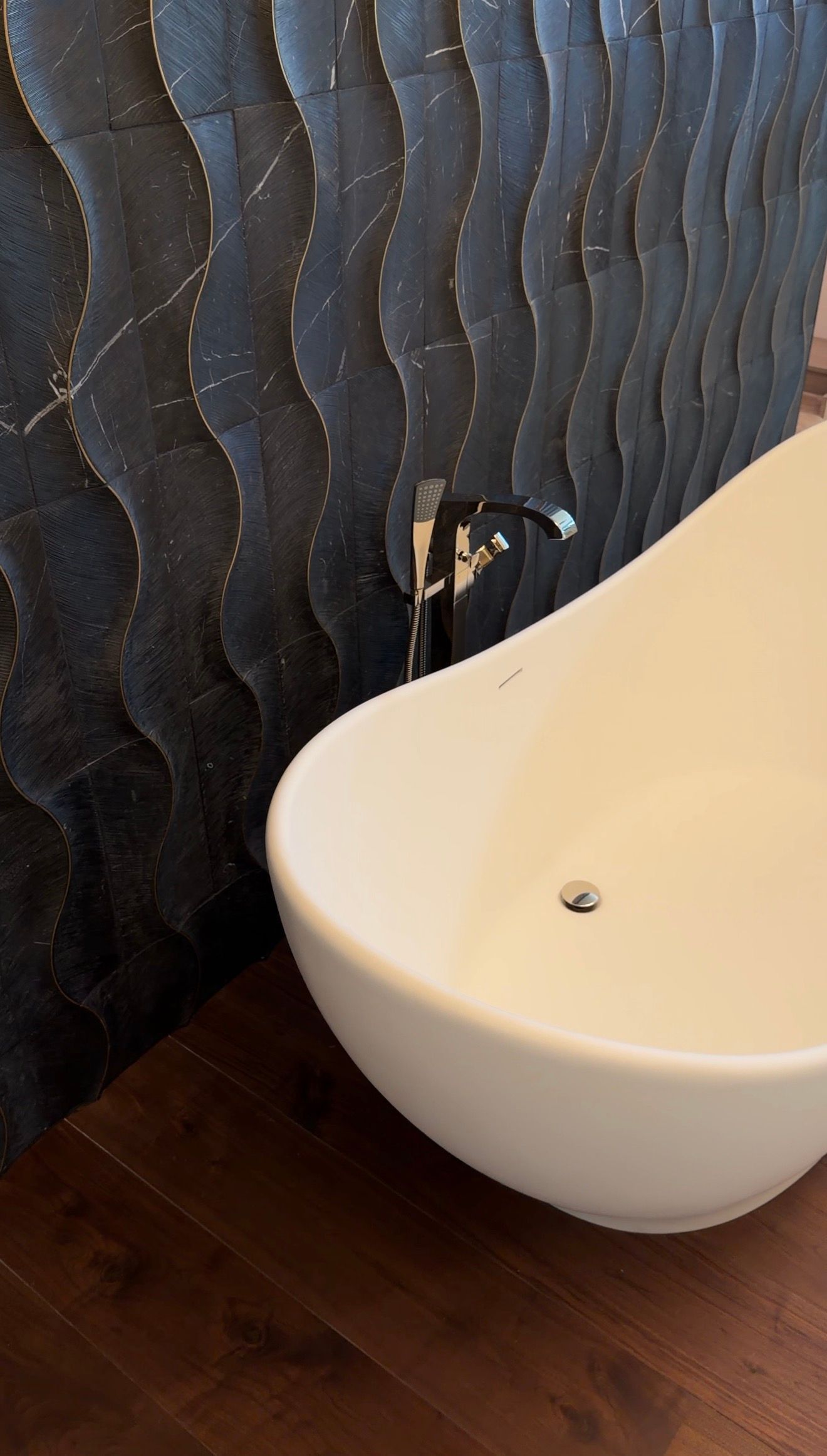 Modern white bathtub beside dark textured tile wall on wood floor