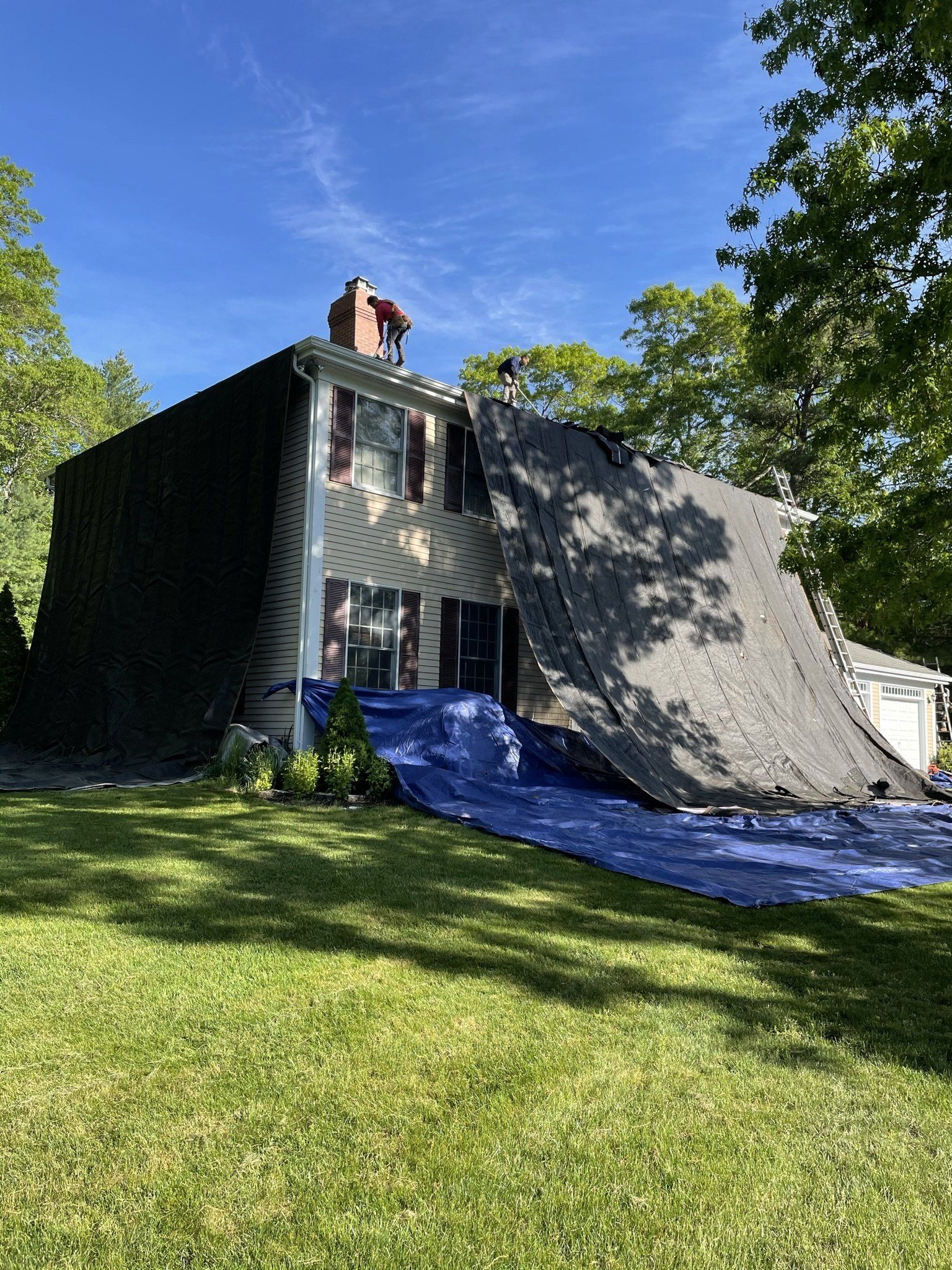 House with damaged roof covered by a black tarp and blue tarp on the ground, grass lawn, sunny day.