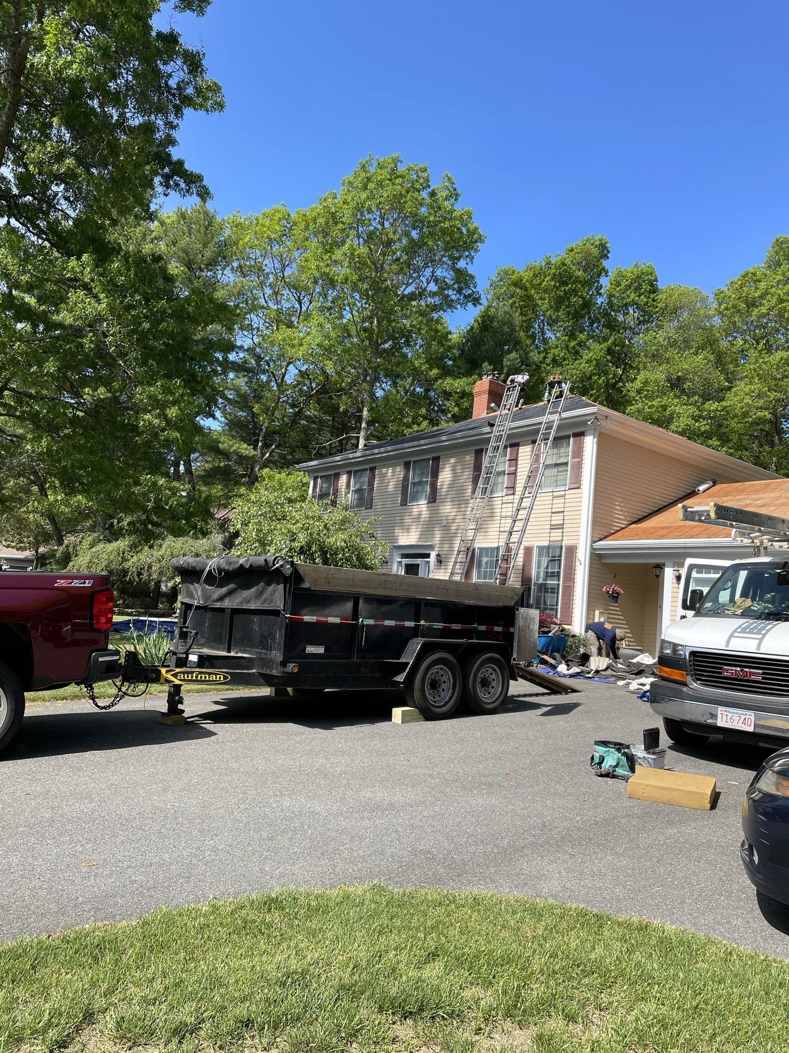 House undergoing roof repair with a black trailer and trucks in the driveway. Workers on a ladder.