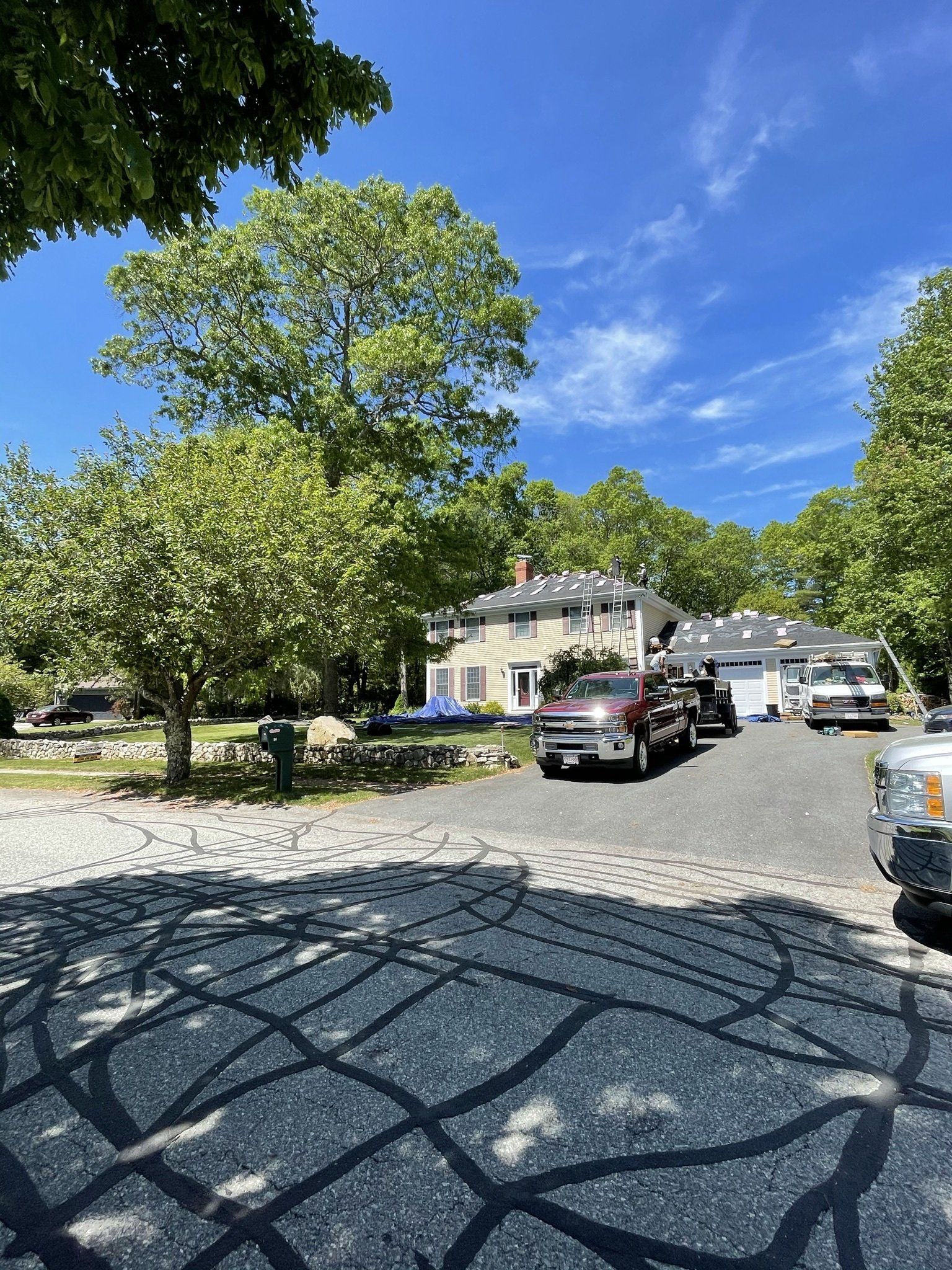 House with vehicles in driveway, sunny day. Trees cast shadows on the gravel, blue sky above.