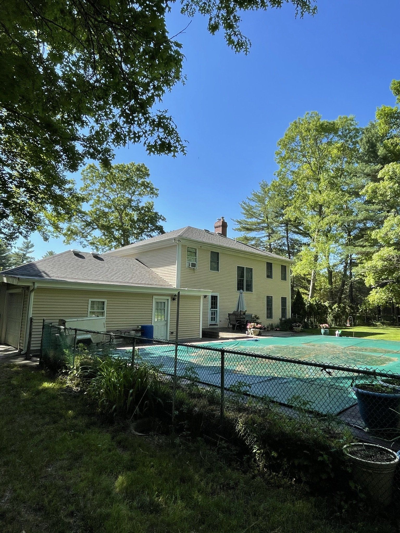 Two-story beige house with a pool covered in green mesh, surrounded by trees and a chain-link fence.