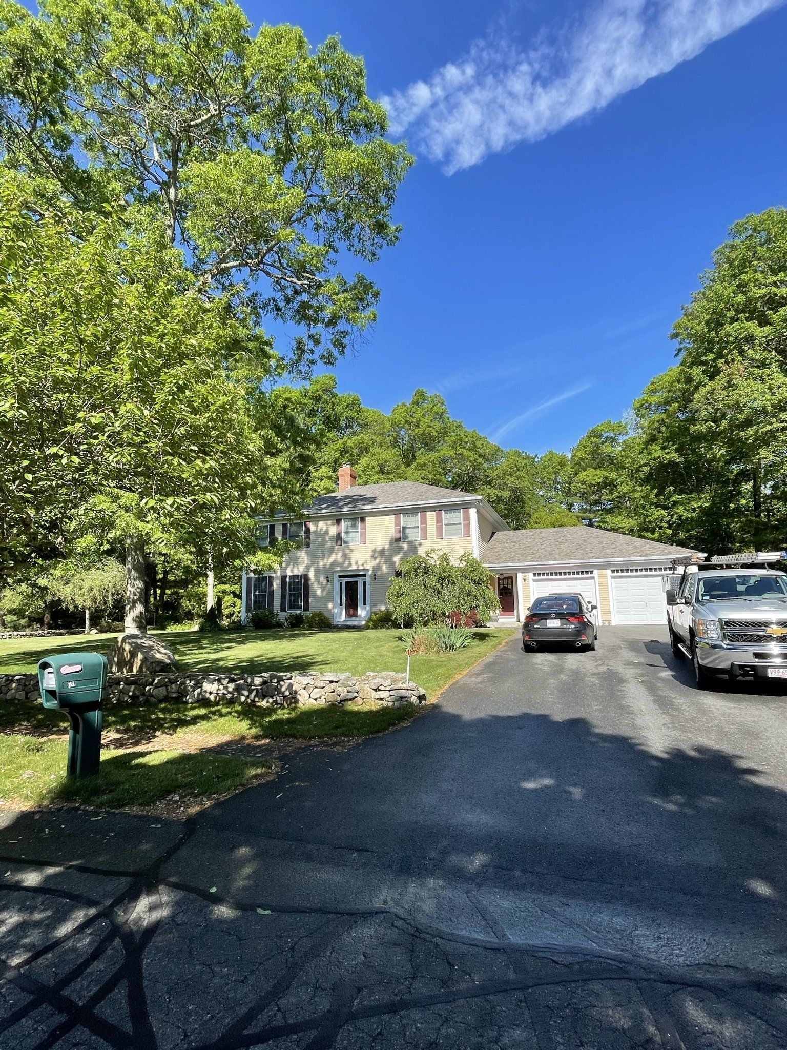 Two-story house with a driveway, trees, and vehicles. Blue sky overhead.
