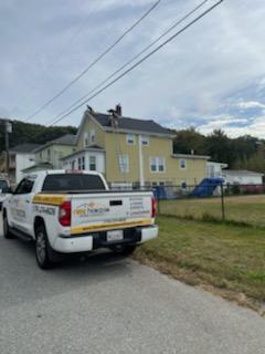 White truck with logo parked near a yellow house with a person on the roof under cloudy sky.