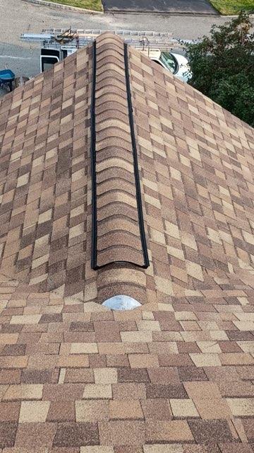 View of a brown shingled roof with a black ridge vent and a chimney.