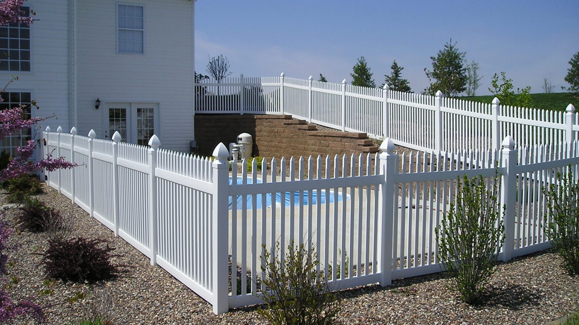 A white picket fence surrounds a swimming pool in front of a house