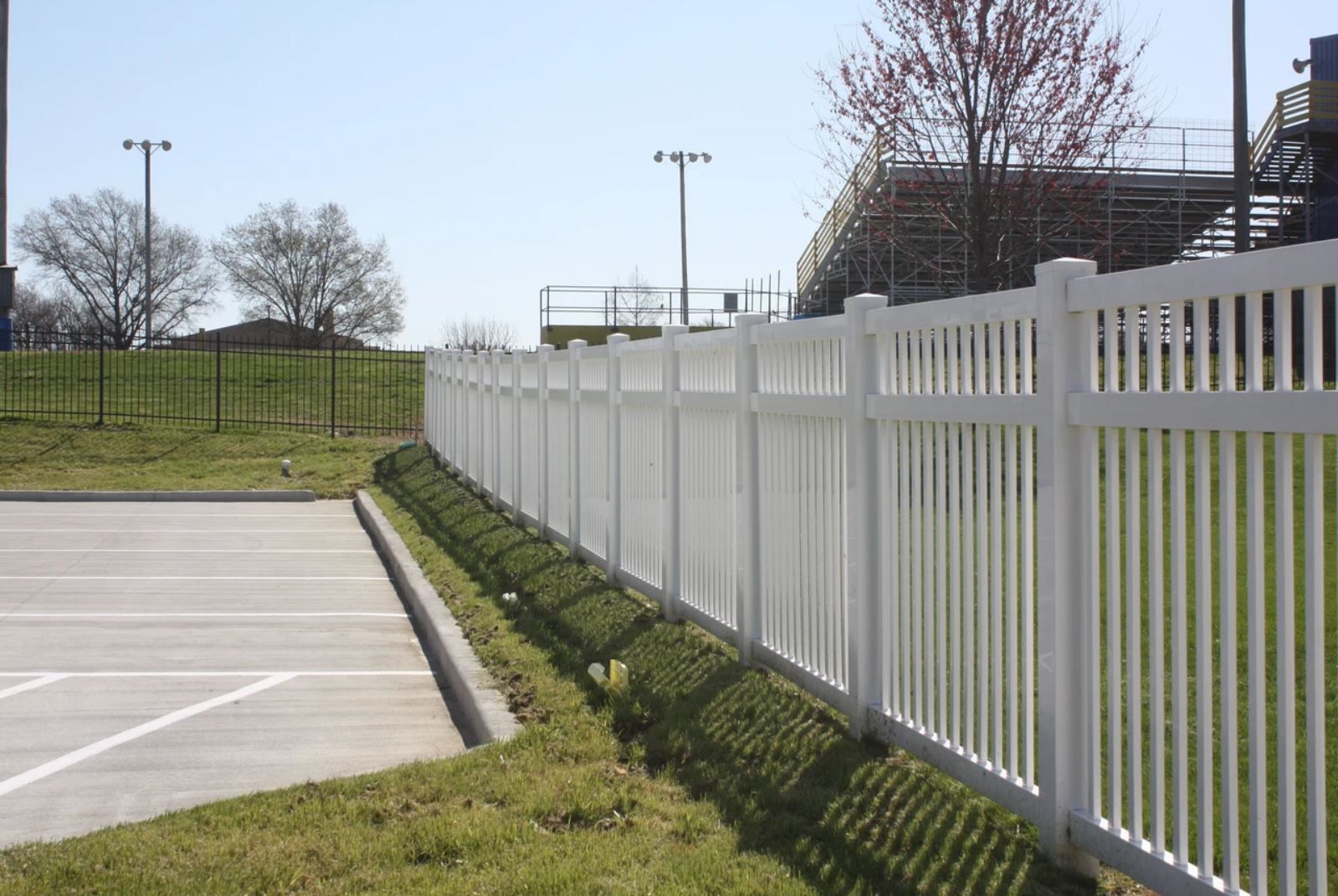 A white fence surrounds a parking lot with a stadium in the background
