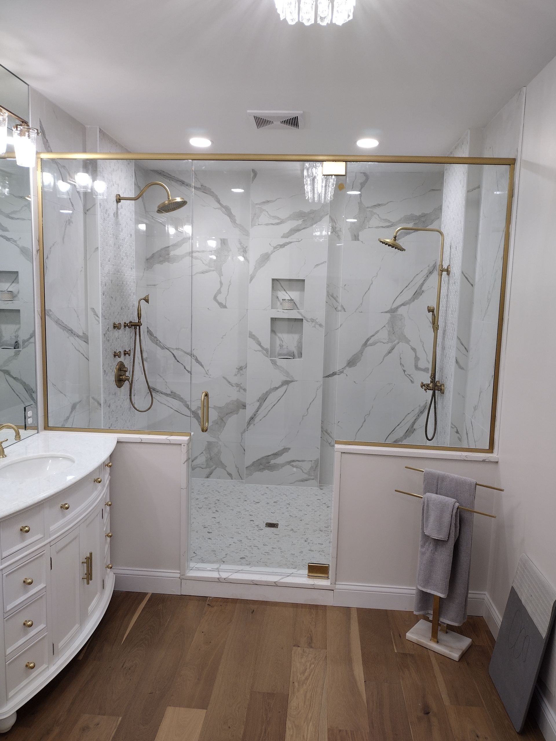 Elegant bathroom with marble-look shower and gold trim. White vanity, wooden floors, and towel rack are present.