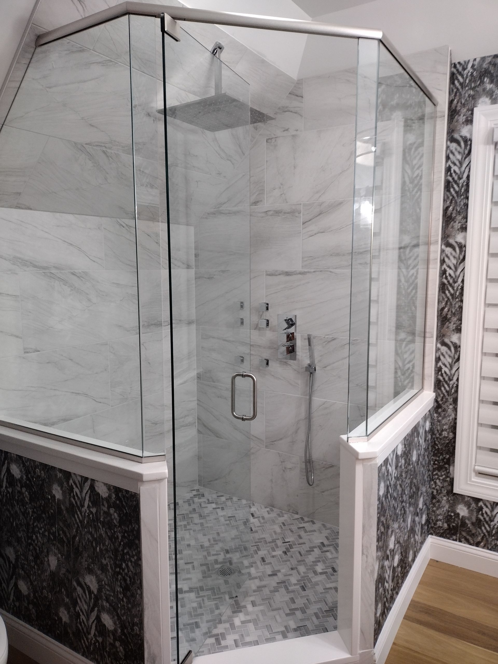 Glass-enclosed shower with white marble walls, pebble floor, and silver fixtures in a bathroom with patterned wall.