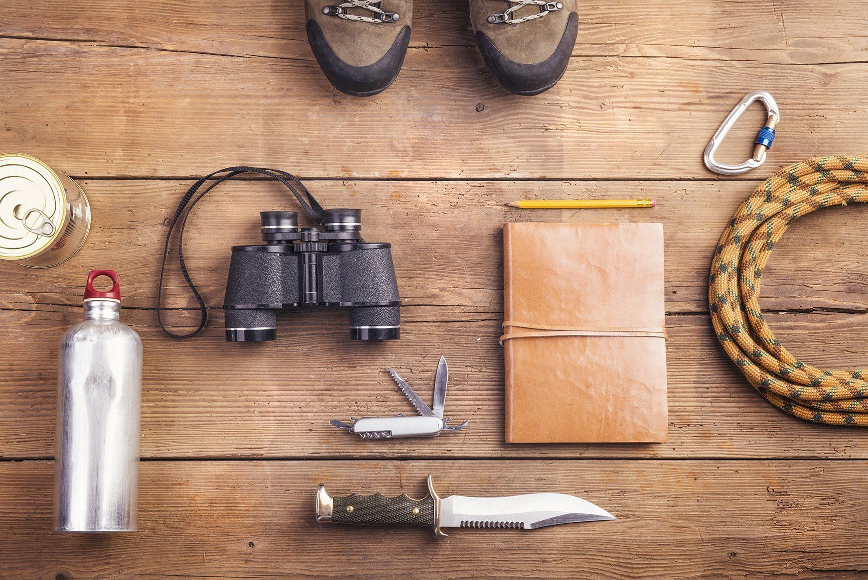 Outdoor gear laid out on wooden planks: binoculars, water bottle, boots, knife, notebook, and coiled rope.