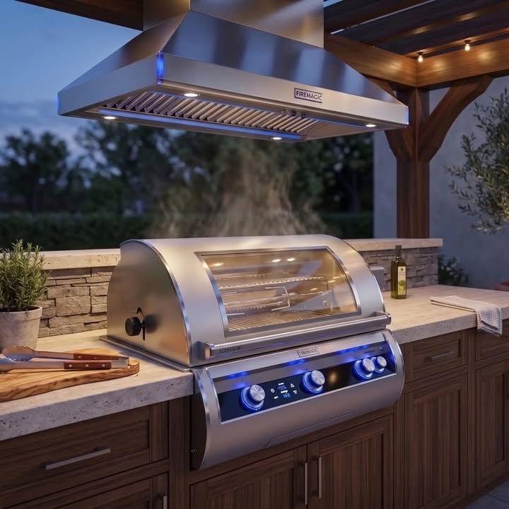 Outdoor stainless steel gas grill with hood under a patio, cooking beside a stone countertop at dusk