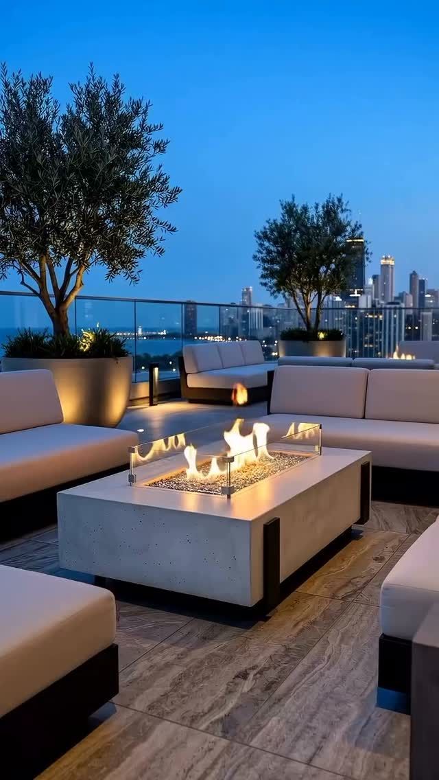 Rooftop lounge at dusk with cushioned seating, lit fire pit, potted trees, and city skyline beyond the water