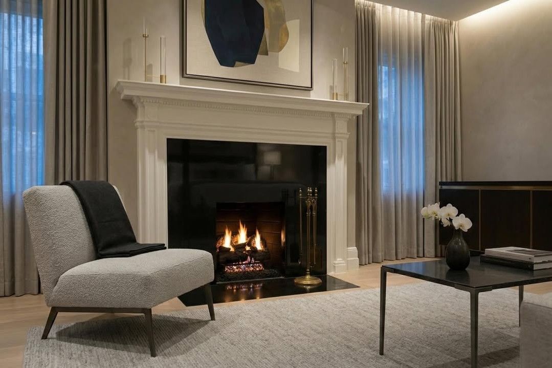 Modern living room with a lit fireplace, gray chair, and coffee table beside tall windows.