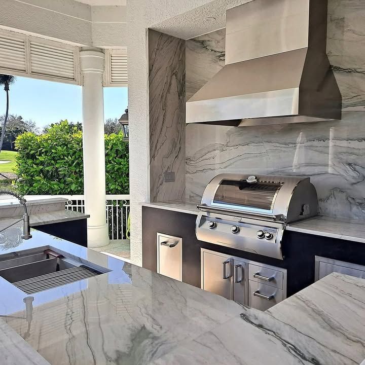Outdoor kitchen with marble countertops, built-in grill, and stainless steel vent hood overlooking a pool and garden.