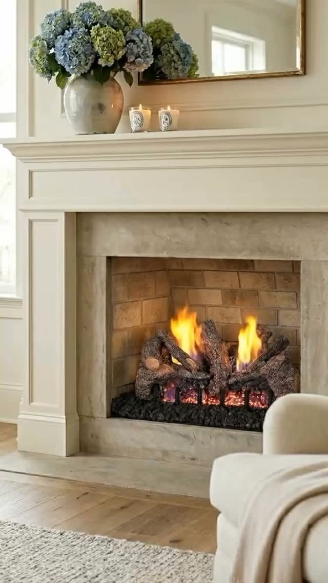 Cozy living room fireplace with a small fire, cream mantel, mirror, and flowers on top.