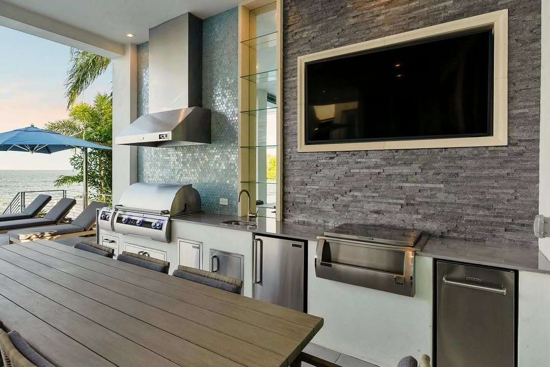 Modern outdoor kitchen with grill, sink, TV, and ocean view beside a wooden table