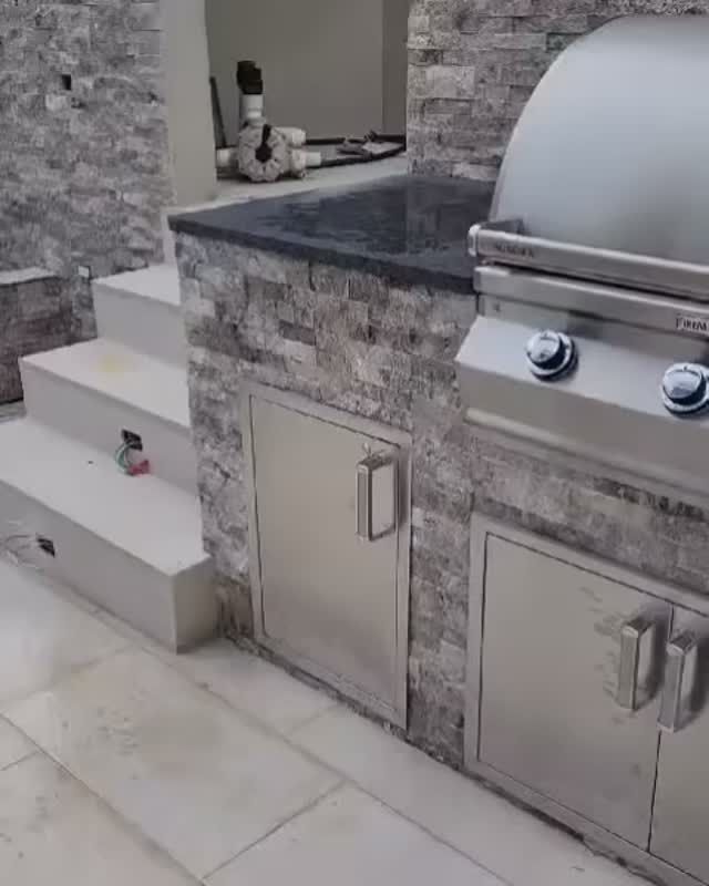 Outdoor stone kitchen with built-in grill, sink, cabinets, and steps.