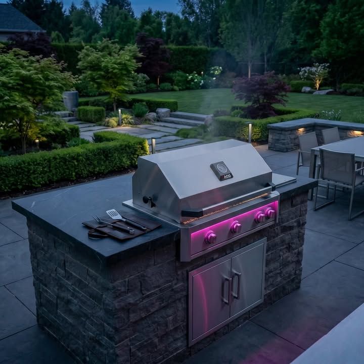 Outdoor patio with a stainless steel grill on a stone island, lit by pink lights at dusk.