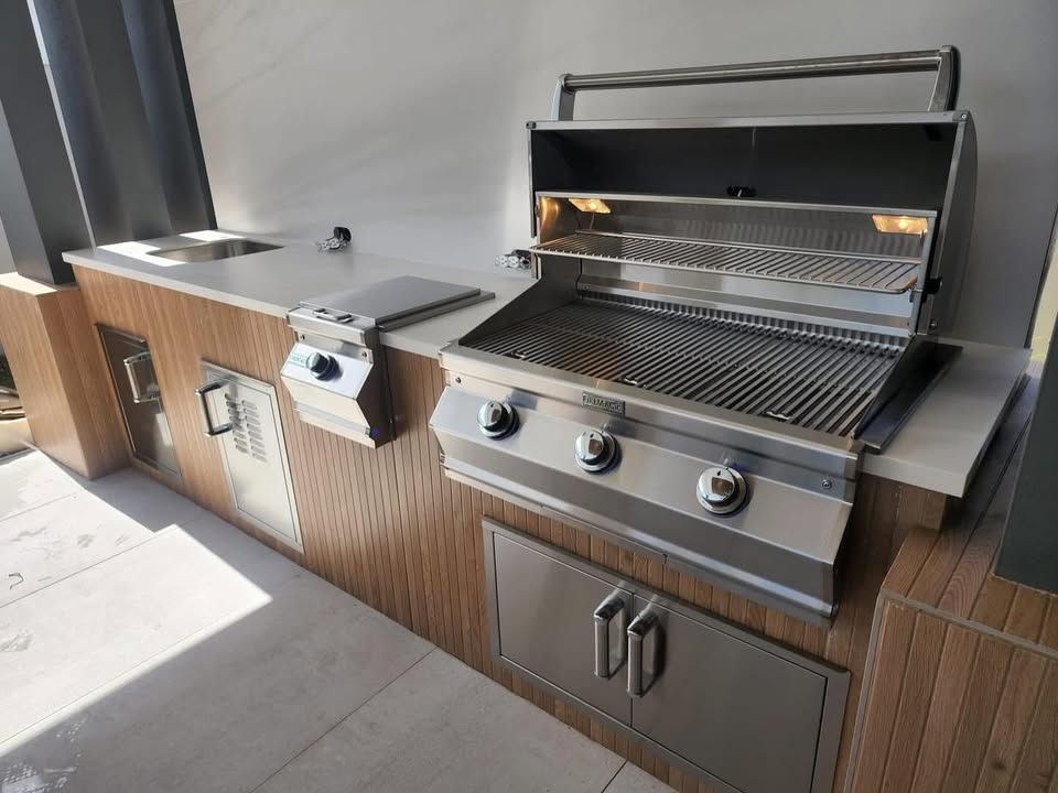 Outdoor stainless steel grill and side burner station with wood-slat cabinetry and sink on a patio