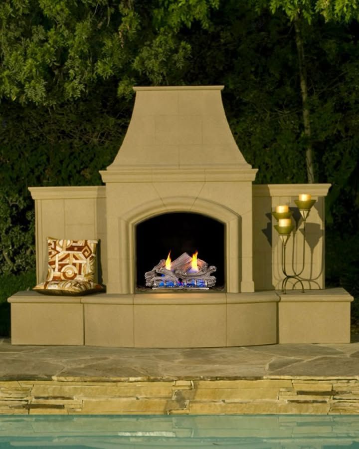 Tan outdoor fireplace with lit fire, built-in bench, and lamp beside a pool at night