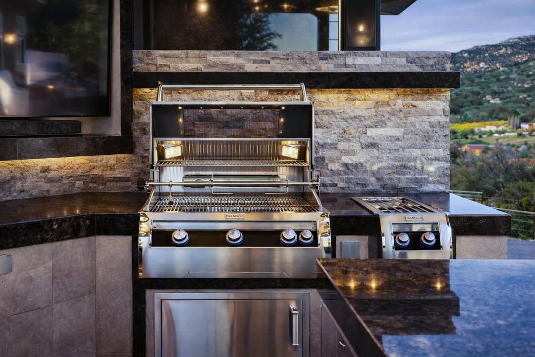 Outdoor built-in stainless steel grill with stone backsplash and dark countertop in a modern patio kitchen