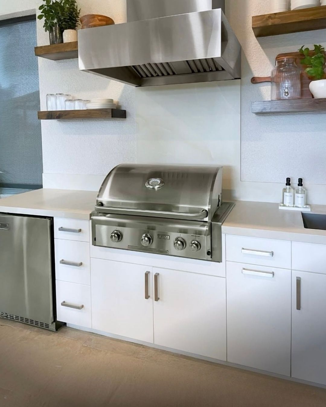 Modern outdoor kitchen with stainless steel grill, white cabinets, and wood shelves on tiled wall