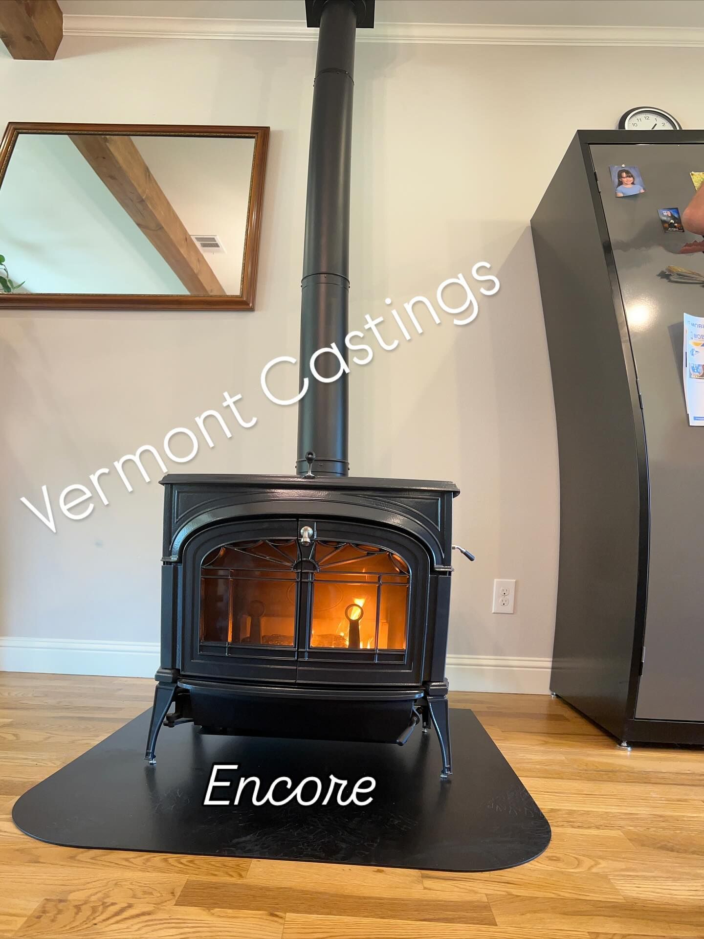 Black Encore wood stove glowing on a black mat in a living room with hardwood floor and mirror wall art