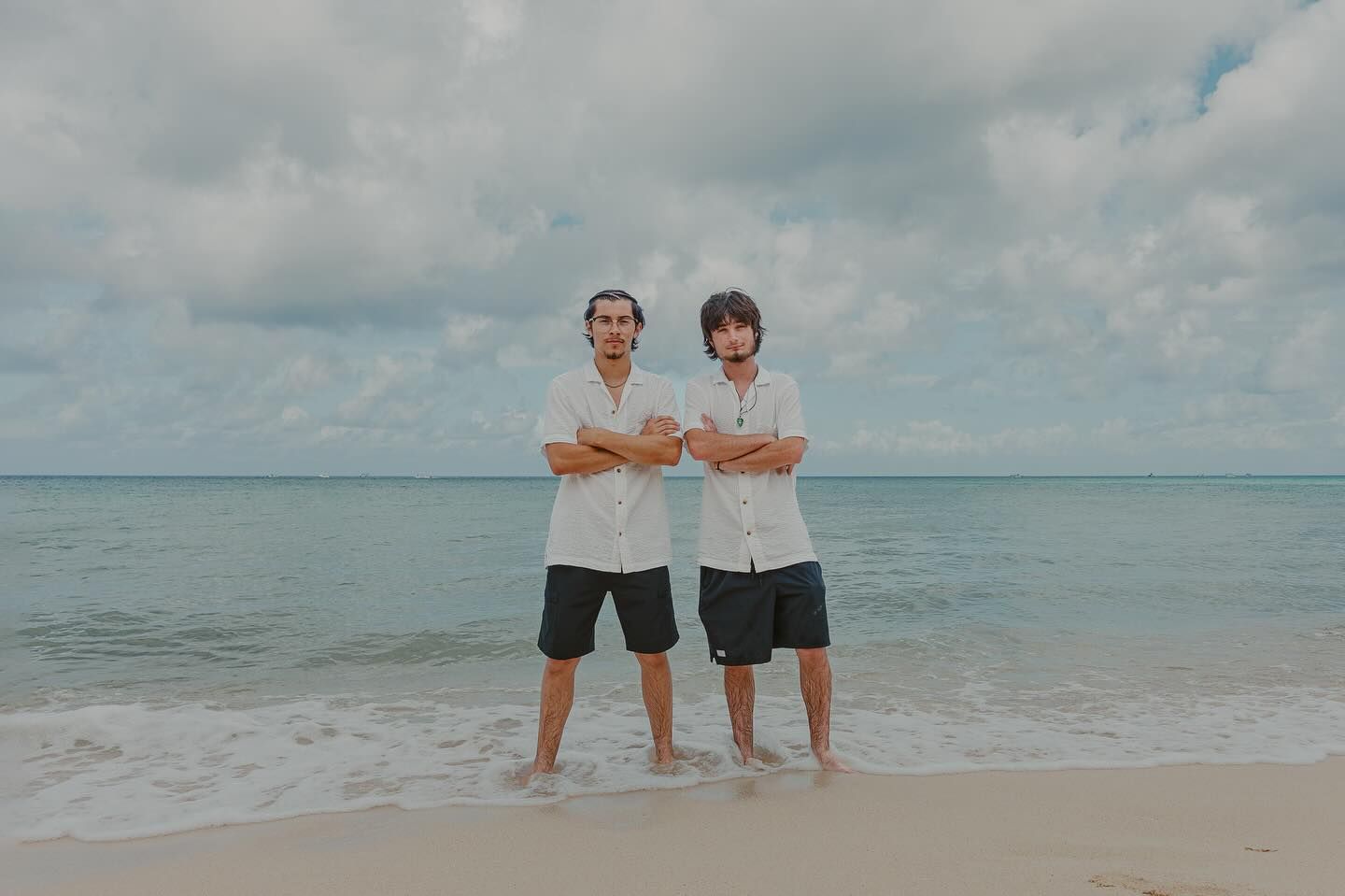 Two people stand on a sandy beach with arms crossed under a cloudy sky.