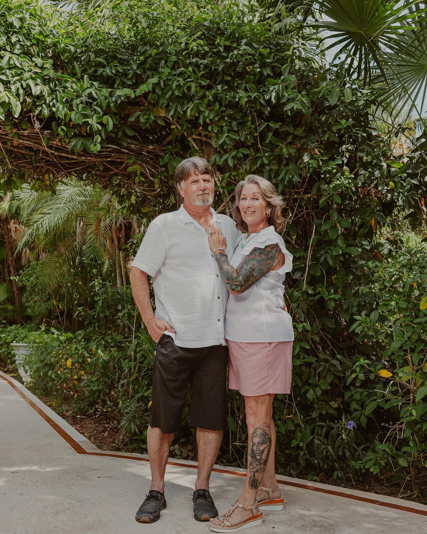 Two people standing together on a sidewalk in front of lush green foliage, smiling and posing for a photo