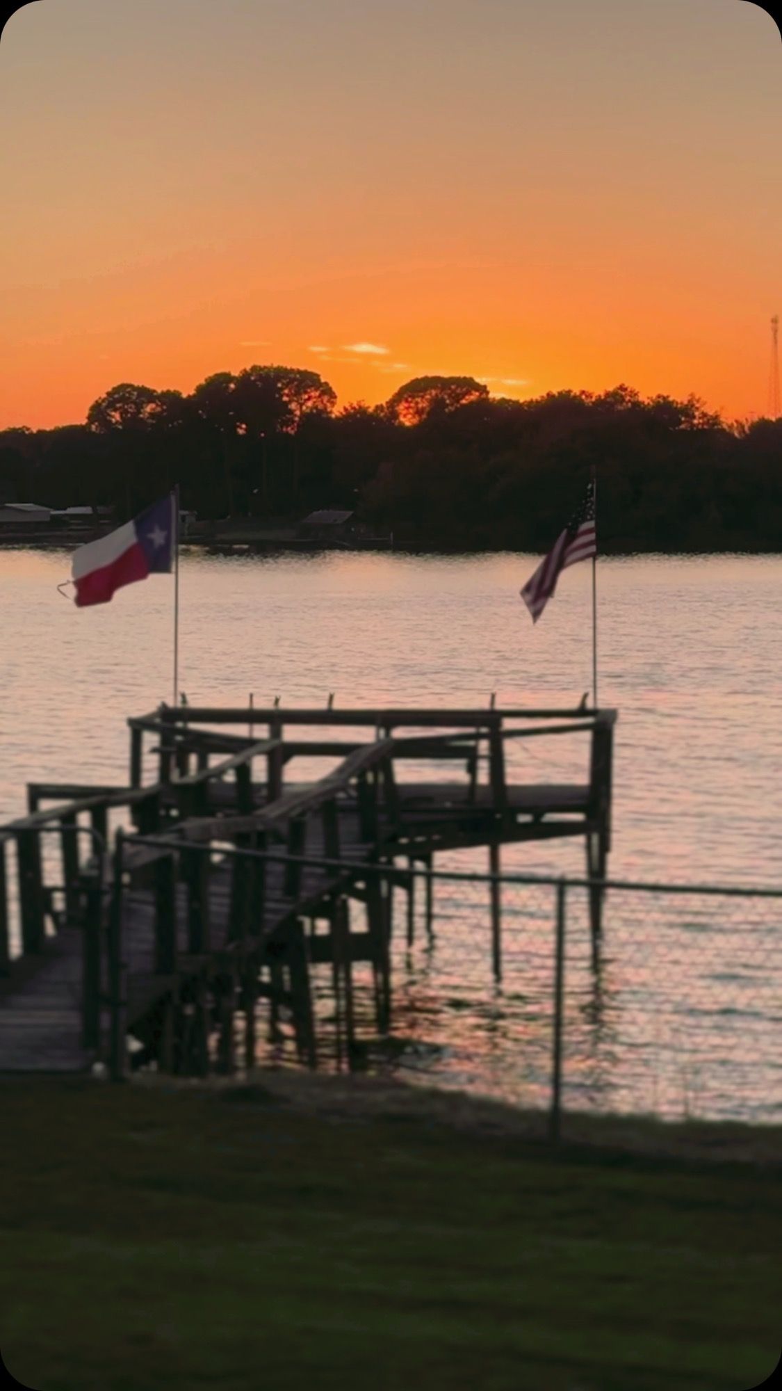 Sunset over a lake with a wooden dock and two flags silhouetted against orange sky